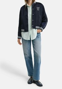 Chaqueta bomber de color azul marino con acentos de franjas blancas, botones de presión en el frente y logo. Combinada con una camiseta de rayas verde claro y jeans azul claro.