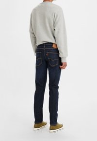 Sudadera gris, jeans azul oscuro con costura amarilla, parche de cuero marrón y zapatillas verdes. Costuras visibles y diseño de bolsillo clásico.