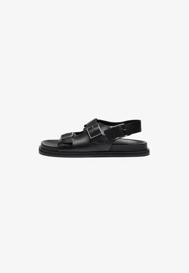 Black leather sandals with a flat sole, featuring two silver buckles and an adjustable heel strap for secure fitting.