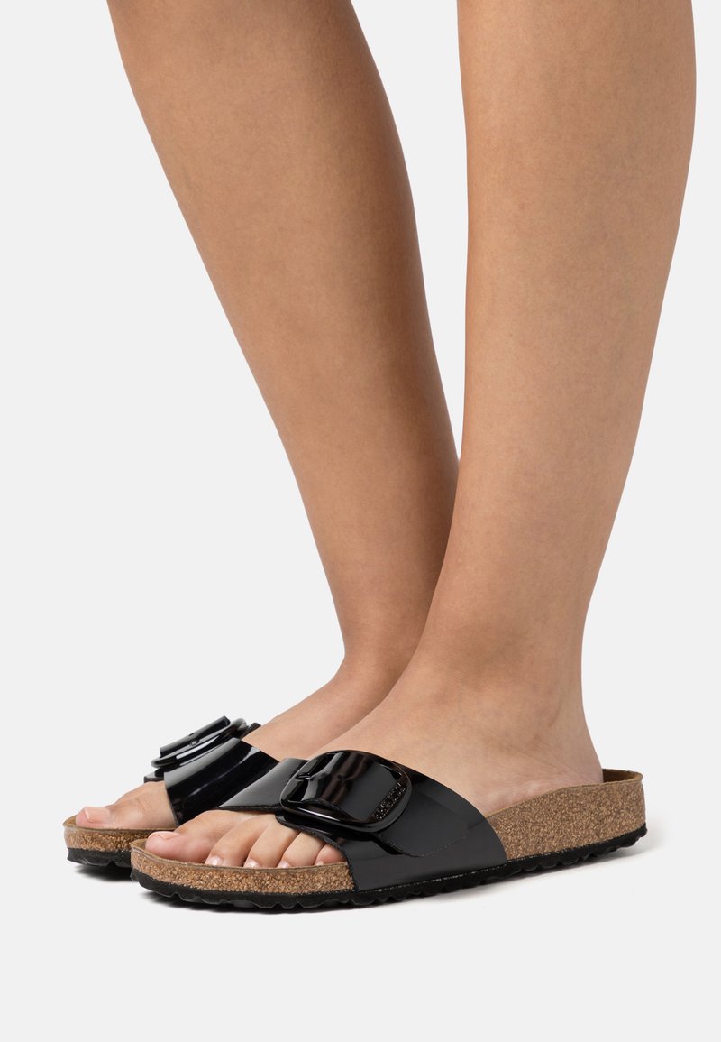 Füße in schwarz glänzenden Riemchensandalen mit großen Schnallen und Kork-Fußbett auf einem weißen Hintergrund.