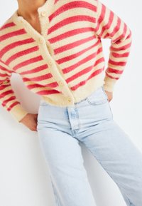 Pletený svetřík s červenými a krémovými vodorovnými pruhy, nadýchaná textura, pět knoflíků a žebrované manžety. Vybaveno s světle modrými džíny.