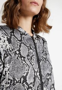 Chaqueta con estampado de serpiente en gris y negro, con acabado texturizado, que presenta un alto cuello y detalle de cremallera visible.