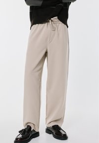 Svijetlosmeđi, široki hlače od mekane tkanine s elastičnim pojasom i vezicom. Uparene s crnim, sjajnim mokasinama s resicama.