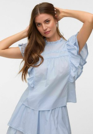 Jeune femme aux longs cheveux bruns portant un chemisier léger bleu à volants et une jupe, posant avec les bras levés derrière la tête sur fond blanc.