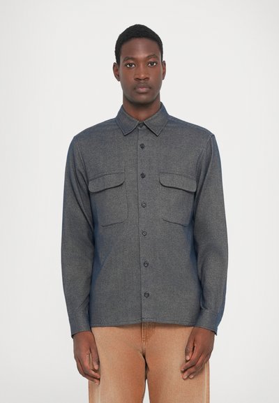 Chemise à manches longues gris foncé avec fermeture par boutons, deux poches poitrine, col soigné et texture lisse. Portée avec un pantalon de couleur claire.