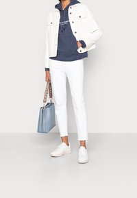 Giacca di denim bianca sopra un hoodie blu navy, jeans bianchi a vita alta, sneakers bianche e una borsa azzurro chiaro con tracolla a catena.