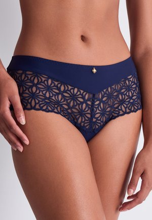 Femme portant un slip en dentelle bleu marine avec des motifs floraux et un petit pendentif doré à la taille, debout devant un fond uni.