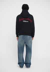 Čierna mikina s kapucňou s vyšitým nápisom "Armani Exchange Milano" v červenej a bielej farbe. V kombinácii s voľnými modrými džínsy a teniskami.