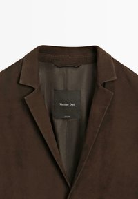 Blazer marrone con una texture in suede liscia, caratterizzato da un colletto a punte e un'etichetta nera all'interno. Design minimalista senza hardware visibili.