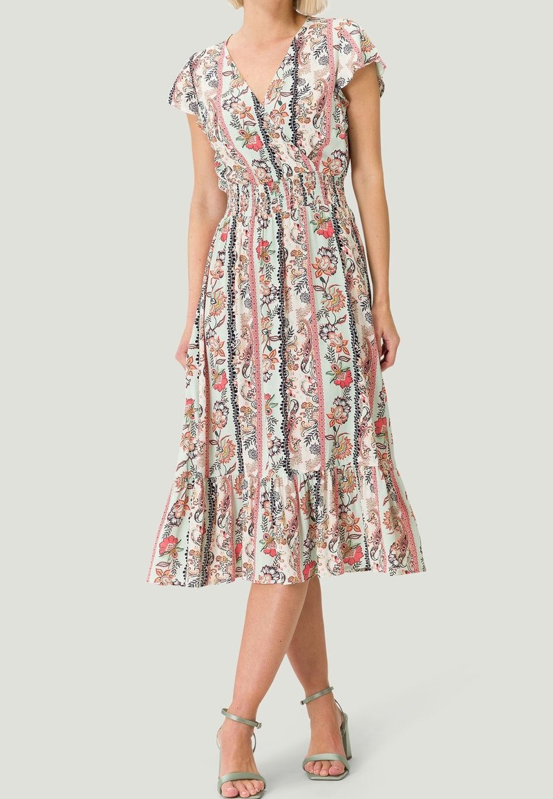 Femme portant une robe midi fleurie à rayures verticales, ourlet volanté et manches courtes à volants, associée à des talons verts à bride cheville.