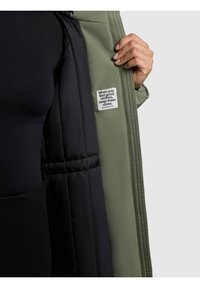 Giacca esterna verde con fodera interna nera. Presenta un'etichetta cucita con del testo. Tessuto morbido e dettagli cuciti all'interno.