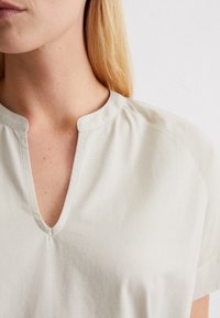 V-kaula kevyesti beige paita, jossa on lyhyet hihat, sileää puuvillakangasta, hienovaraisia rypytystä kaula-aukossa ja siisti, yksinkertainen muotoilu.