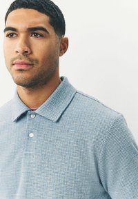 Lyseblåt tekstureret polo-shirt med krave og to knapper, der har et ternet mønster og en blød stoffinish.
