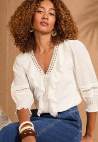 Blusa bianca con bordo arricciato, dettagli in pizzo e scollatura a V profonda. Presenta maniche a palloncino e tessuto testurizzato. Abbinata a jeans e bracciali in legno.