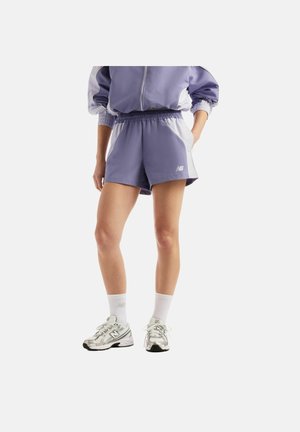 Person iført lilla og hvide atletikshorts, en matchende jakke, hvide New Balance sokker og sølvfarvede sneakers mod en ensfarvet baggrund.