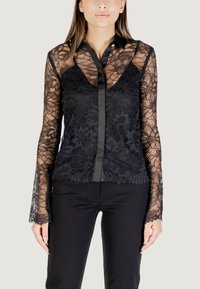 Blusa nera in pizzo con maniche lunghe, caratterizzata da una patta in raso e motivi floreali, abbinata a pantaloni neri aderenti.