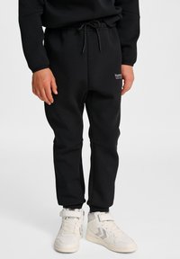 Zwarte sweatpants met een trekkoord in de taille, zijzakken en een klein logo op de heup, gecombineerd met witte high-top sneakers.