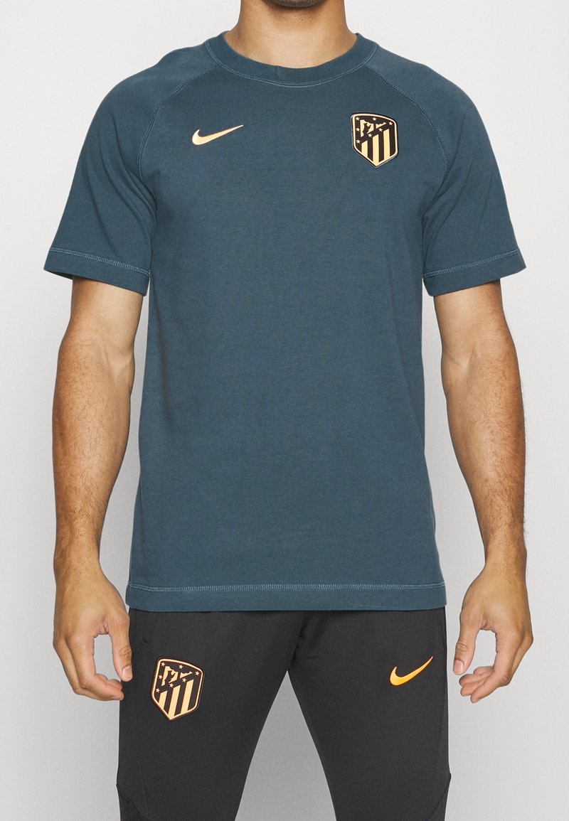 Torso masculino que lleva una camisa de manga corta azul grisácea y pantalones cortos negros con logotipos de Nike en naranja y escudos del club de fútbol Atlético de Madrid.