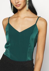 Femme portant un caraco en satin vert avec de fines bretelles et de longues boucles d'oreilles pendantes perlées sur fond blanc.