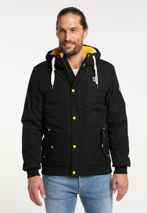 Mann mit Bart, der eine schwarze Kapuzenjacke mit gelben Knöpfen und Futter trägt, kombiniert mit hellblauen Jeans, stehend vor einfachem Hintergrund.