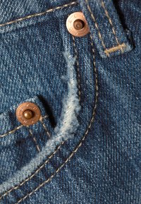 Κοντινή λήψη μπλε υφάσματος τζιν με φθαρμένη άκρη και χαλκομανίες σε ραφή τσέπης.