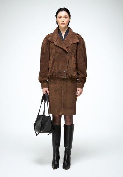 Vrouw die een oversized bruine suède jas en rok draagt, zwarte maillots, zwarte laarzen tot de knie en een zwarte leren handtas vasthoudt.