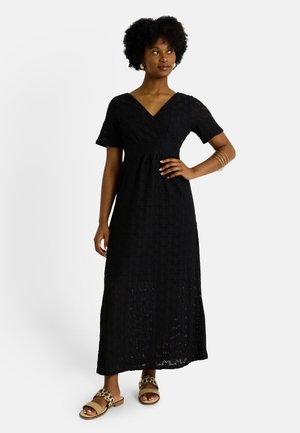 Femme debout avec une main sur la hanche, portant une robe maxi en dentelle noire, des bracelets en or et des sandales à imprimé léopard sur un fond blanc.