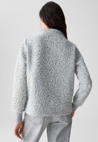 Grauer strukturierter Pullover mit hohem Kragen, weichem, flauschigem Stoff und gerippten Bündchen. Die Rückansicht zeigt eine kastige Passform und subtile Farbvariationen.