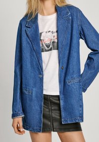 Modrý denimový blejzr s klopovým límcem a uzávěrem na knoflíky; spárovaný s bílým tričkem s grafikou a černou koženou mini sukní.