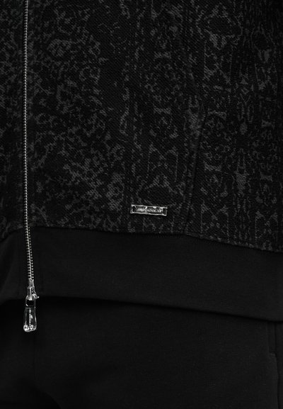 Prim-plan al unei jachete negre cu textură și modele întunecate subtile, fermoar argintiu și o etichetă mică dreptunghiulară din metal cu brand pe tivul elastic.