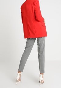 Blazer rouge sur mesure à manches trois-quarts, pantalon gris à carreaux, et escarpins blancs à bout ouvert, vus de dos.