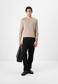 Pull en V beige, jeans noirs, sac en cuir noir et chaussures noires. Texture lisse avec des détails de design subtils, y compris un petit logo.