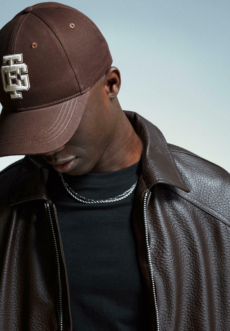 Brown cap with logo, black T-shirt, silver chain necklace, and a dark brown leather jacket with textured finish, captured in a downward gaze.