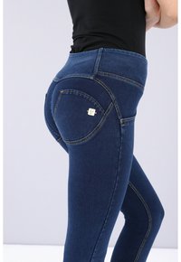 Pantalones vaqueros de mezclilla azul oscuro con tiro alto, con costuras doradas, bolsillos traseros curvados y un pequeño acento de marca en el bolsillo izquierdo.