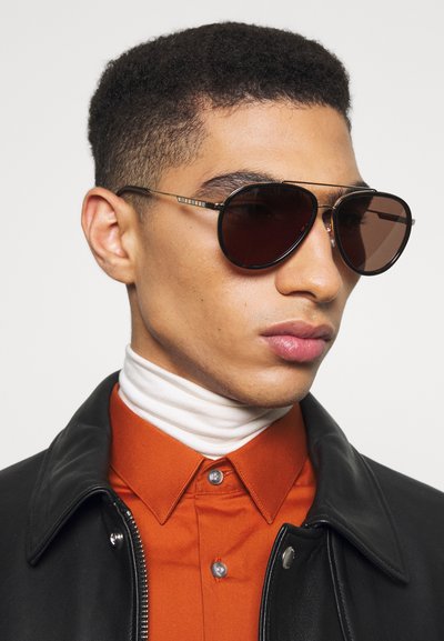 Solsikker med mørkt tonede linser og slanke sorte metalrammer. Modellen har en sort læderjakke og en orange skjorte med knapper på, samt en hvid turtleneck.