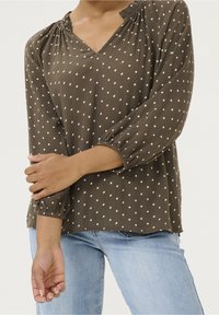 Blouse vert olive à pois blancs, col en V, manches longues bouffantes, détails froncés au niveau du col et des poignets. Tissu texturé.