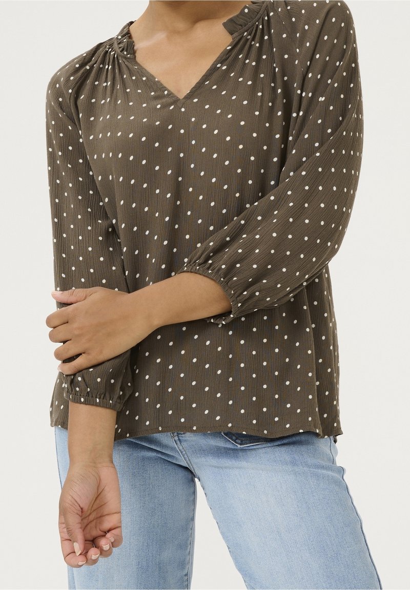 Blouse vert olive à pois blancs, col en V, manches longues bouffantes, détails froncés au niveau du col et des poignets. Tissu texturé.