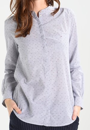 Frau trägt ein hellblaues, langärmliges Hemd mit kleinen roten und blauen Punkten, Hände in den Taschen, vor einem einfarbigen Hintergrund.