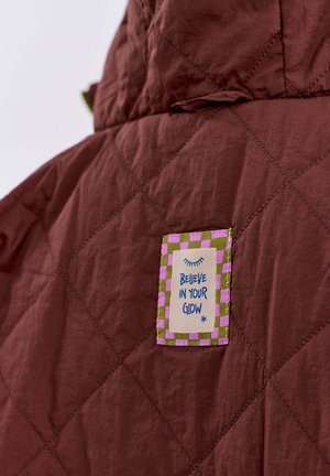 Prošívaná hnědá bunda s mnohobarevným nápletem s kostkovaným okrajem a textem "VĚŘTE VE SVŮJ LESK."