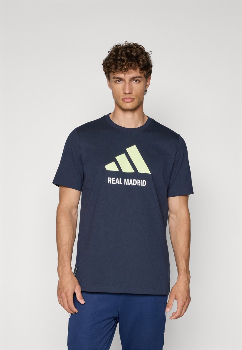 Marineblå bomulds T-shirt med et lysgrønt grafisk design med tre skrå striber og teksten "REAL MADRID", rund halsudskæring og korte ærmer.
