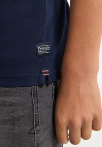 Námořnický modrý polo trikotu s texturovaným vzorem, se tkanou nášivkou "Petrol Ind" a růžovými švy na lemu. Černé džínové kalhoty.