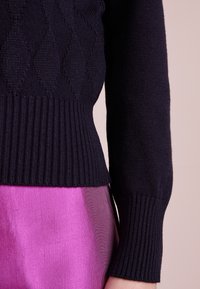 Navy knitted sweater with diamond pattern, ribbed hem and cuffs. Soft texture and fitted design, paired with a vibrant pink skirt.