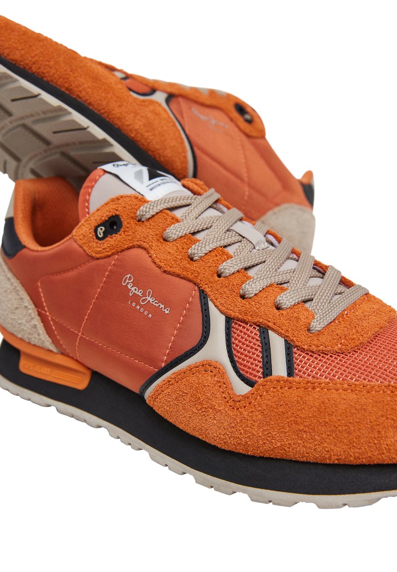 Zapatillas naranjas con materiales de ante y malla, que presentan acentos en beige, una suela con diseño y cordones grises. Logo visible en el lateral.