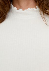 Bílý žebrovaný top s vysokým výstřihem a volánkovým okrajem. Tkanina vypadá měkce a lehce, čímž podtrhuje jednoduchý design.