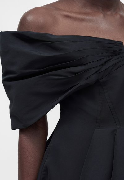 Άτομο με σκουρόχρωμο δέρμα που φοράει μαύρο φόρεμα με ακάλυπτους ώμους και μεγάλο, διπλωμένο φουσκωτό μανίκι σε ένα χέρι, σε απλό φόντο.