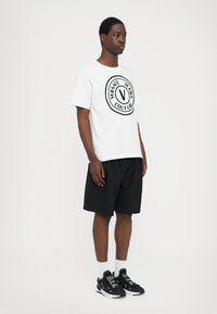 Balts kokvilnas T-krekls ar melnu aplokšņu logotipu "VERSACE JEANS COUTURE" priekšpusē, kombinēts ar melnām šortiem un melniem sporta apaviem.
