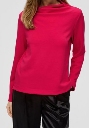 Kobieta w jasnoróżowej bluzce z długim rękawem i dekoltem w łódkę oraz czarnych satynowych spodniach na szarym tle.