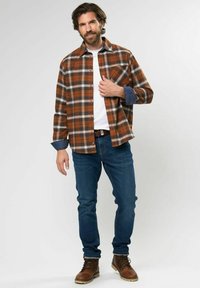 Brązowa i biała koszula flanelowa w kratę z długimi rękawami, akcenty z niebieskiego denimu, pod nią biała koszulka, niebieskie dżinsy i brązowe buty.