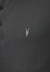 Dunkelgraues Poloshirt aus strukturiertem Stoff, mit einer Knopfleiste mit drei Knöpfen und einem kleinen gestickten Ziegenschädel-Logo auf der Brust.