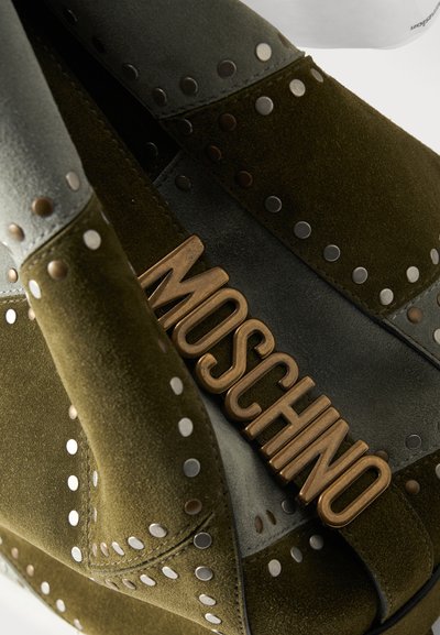 Σκούρο πράσινο σουέντ παπούτσι με ασημένιες καρφίτσες και μεταλλική πλακέτα με το λογότυπο "MOSCHINO" στο λουράκι, που δείχνει λεπτομερή υφή και σχέδιο.